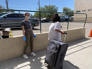 A student smiles for the camera. She is wearing a mask. To the right is a person pulling a suitcase with food. The wonderful students at Bosque School are giving back in many ways.